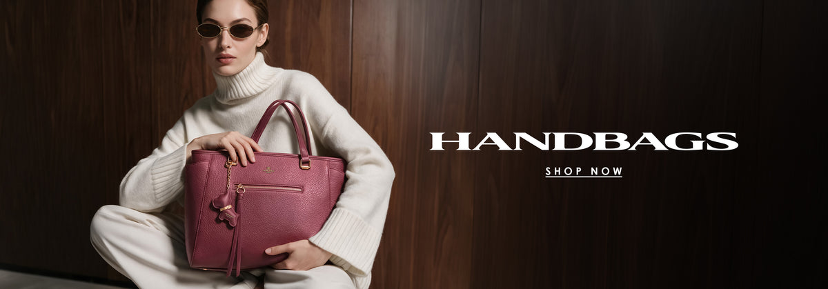 A woman in a white knit turtleneck sweater and sunglasses sits holding a dusty rose leather handbag against a dark wood-paneled background, with the text 'HANDBAGS SHOP NOW' on the right.